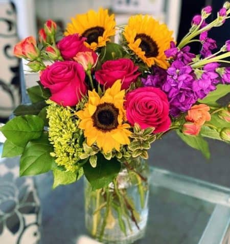 A vibrant assortment of sunflowers, purple stock, hot pink roses, and orange spray roses designed in a glass clear glass vase is sure to put a smile on anyone's face.